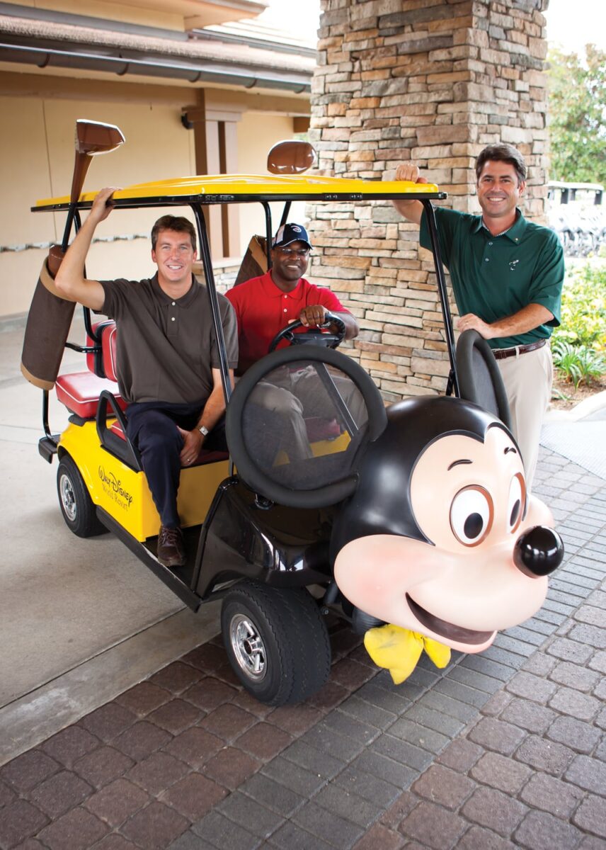 Three men smiling with a yellow golf cart featuring a large Mickey Mouse face on the front.