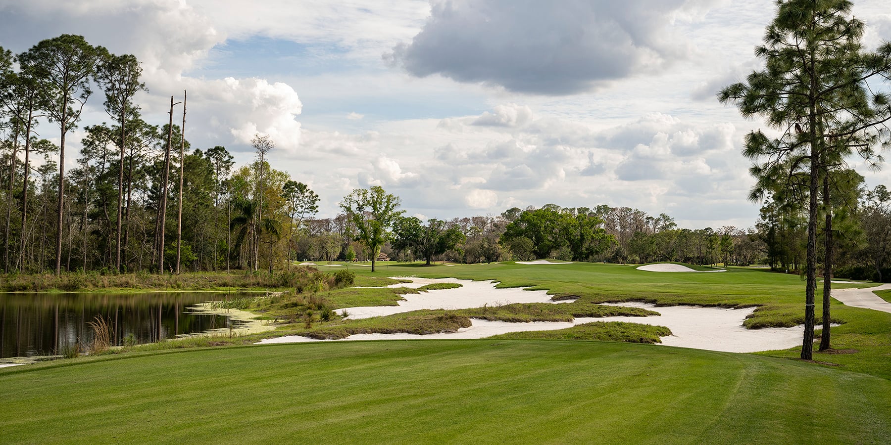 A golf course with sand bunkers, a pond, trees, and a cloudy sky in the background.