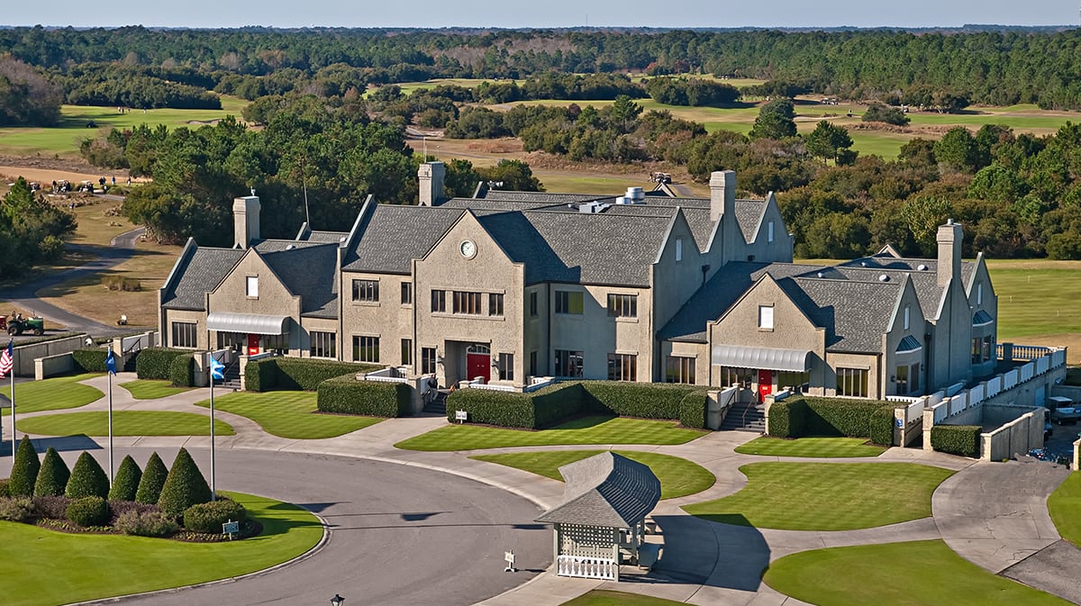 Large country club building with manicured lawns and trees, surrounded by golf course and driveways.