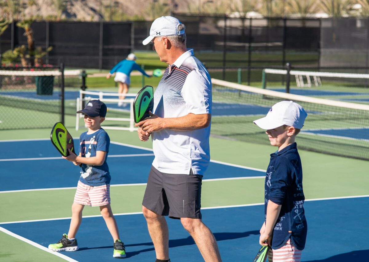 An adult teaching two young boys how to play pickleball on an outdoor court.