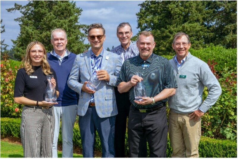 Six people standing outdoors, holding glass awards, smiling with trees and greenery in the background.