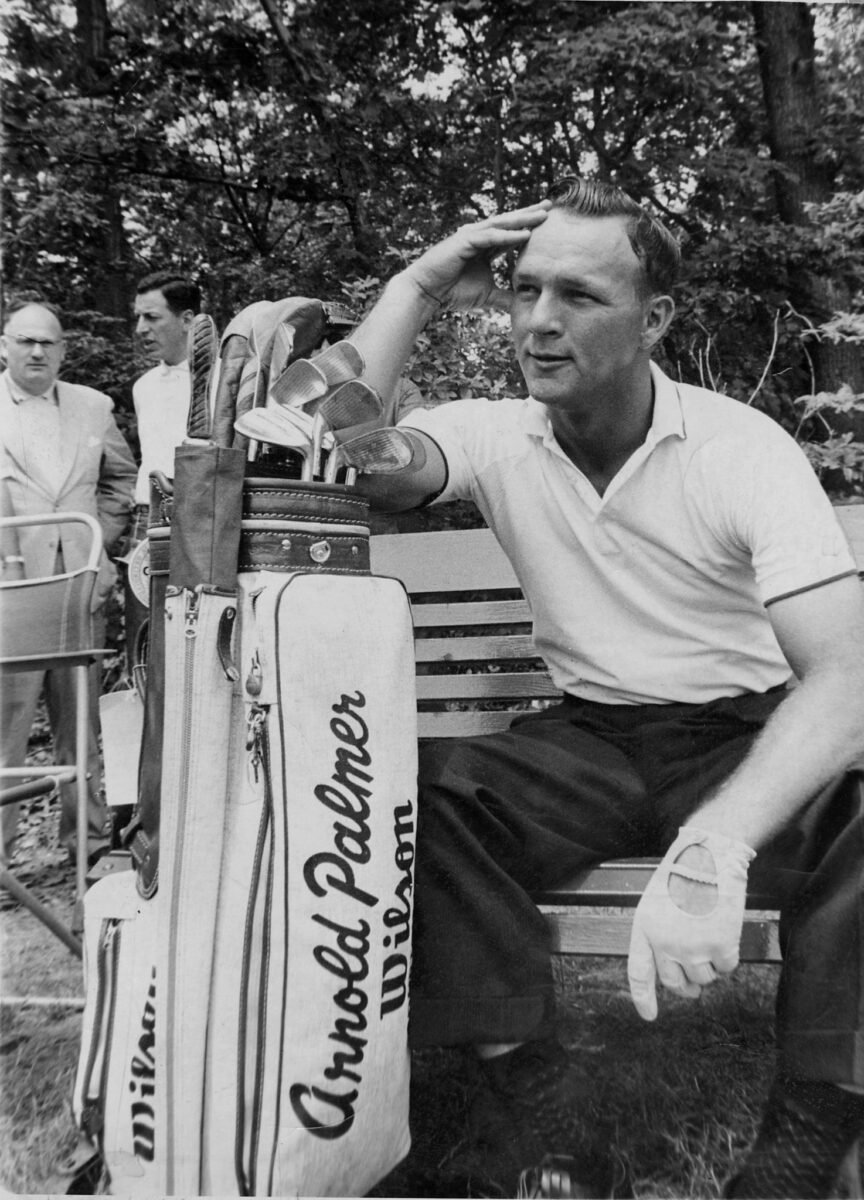 A golfer sits on a bench beside a golf bag labeled Arnold Palmer Wilson, raising his hand to his brow.