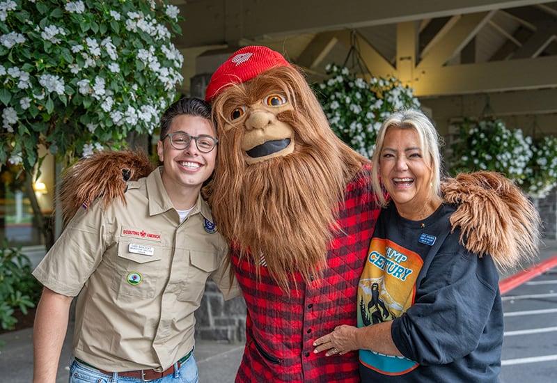 Three people, including one in a Bigfoot costume, smile and pose together outdoors near hanging plants.