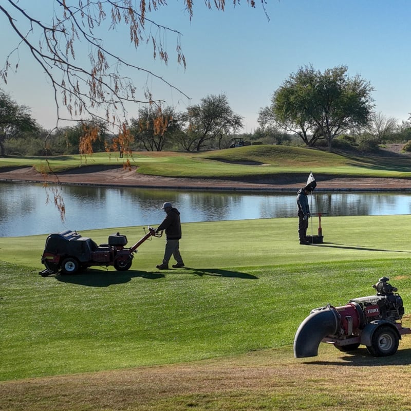 Two workers maintain a golf course near a pond, using lawn equipment on a sunny day.