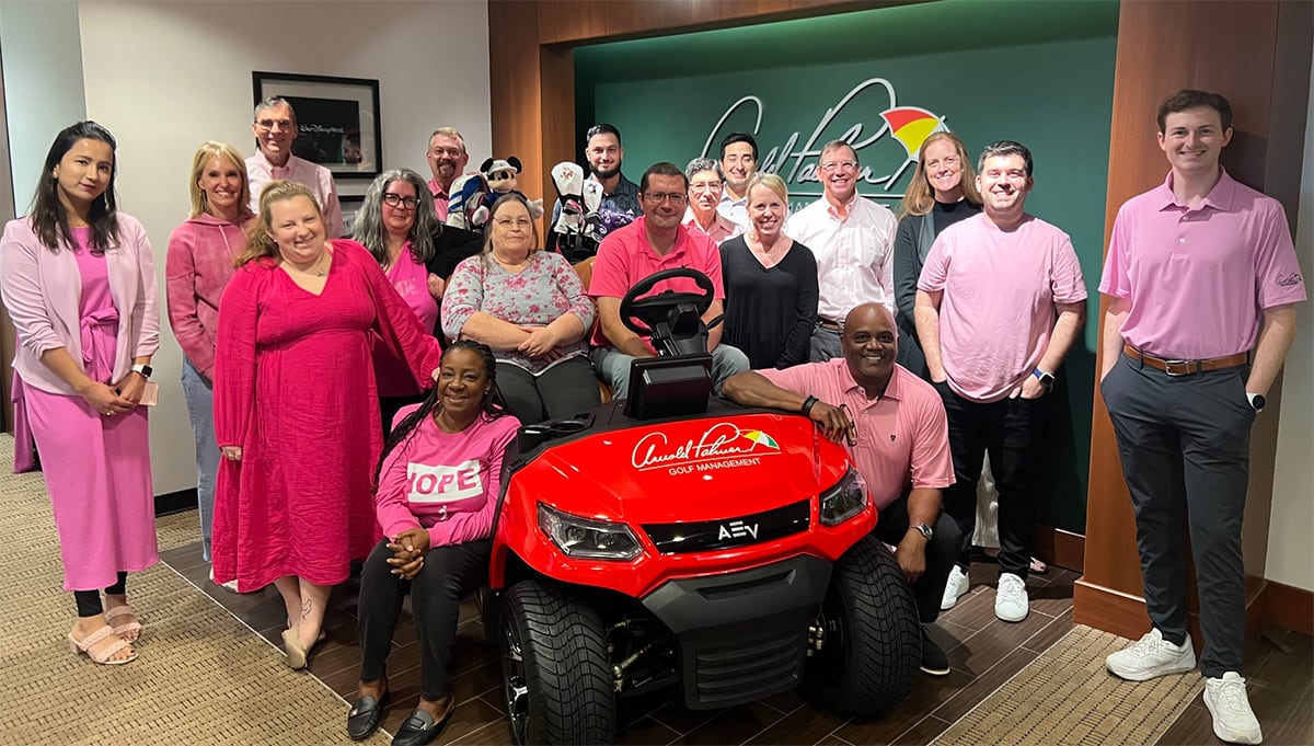 Group of people wearing pink pose around a red golf cart inside an office with a green and brown background.