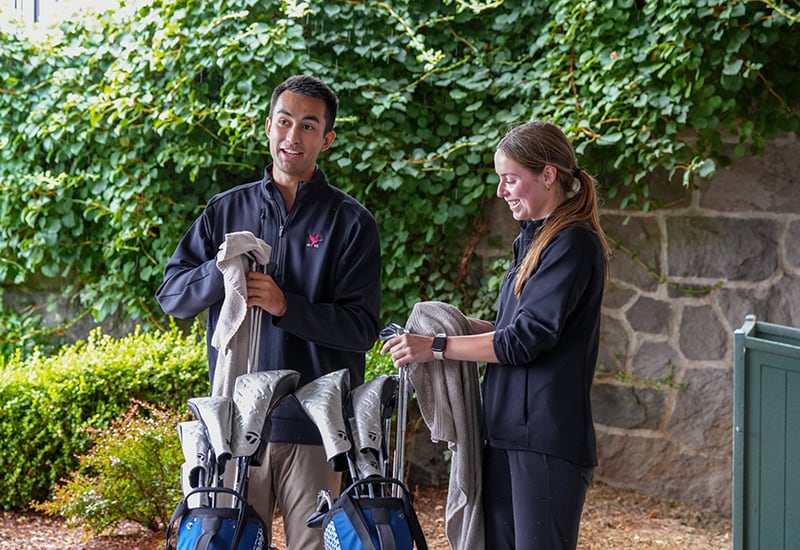 Two people smiling while cleaning golf clubs near a stone wall and green foliage.