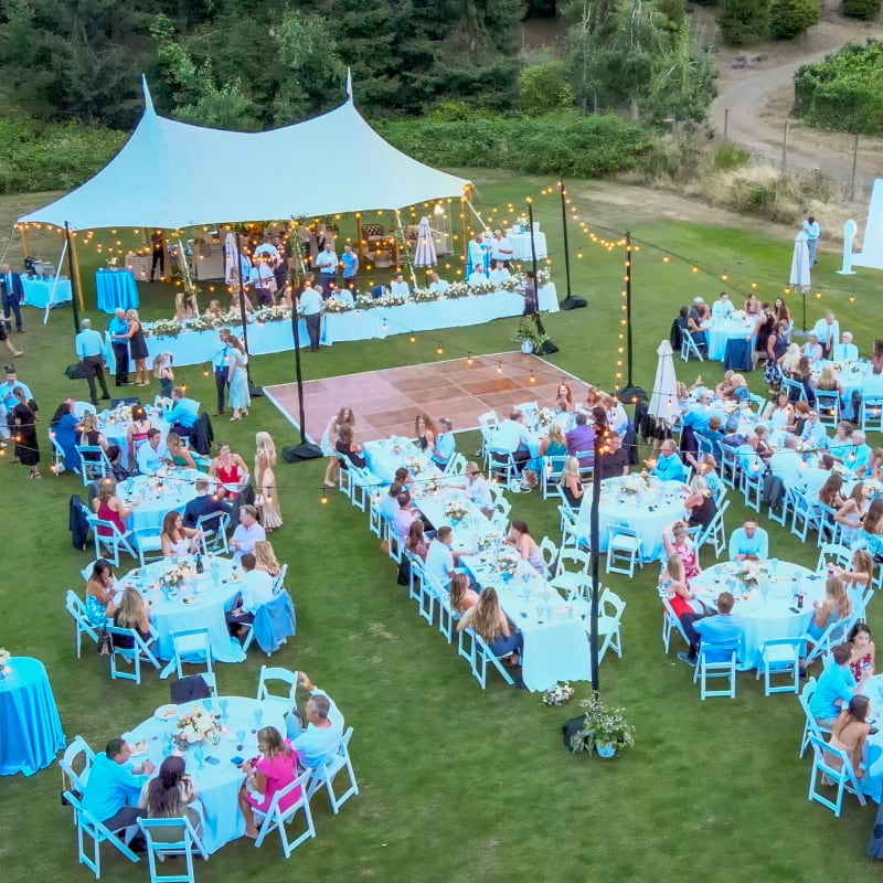 Outdoor wedding reception with round tables, guests dining, and a band playing under a white tent on a lawn.