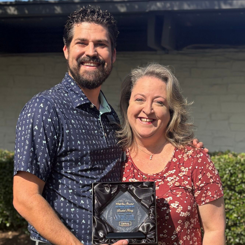 Two smiling people stand outside, holding a black award plaque together in front of green bushes.