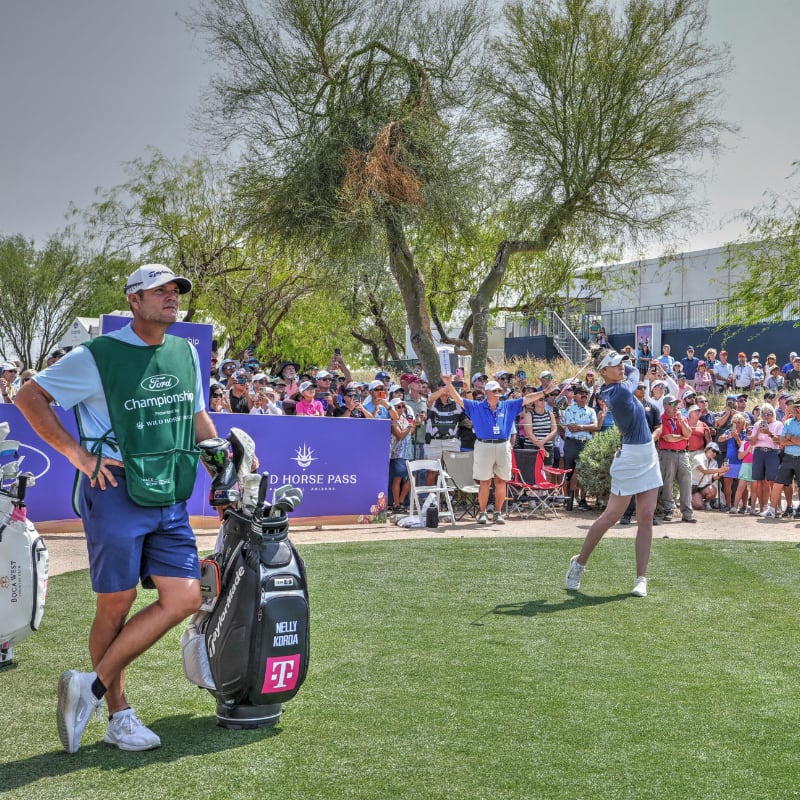 A golfer swings a club while a caddie stands nearby; a crowd watches at an outdoor golf tournament.