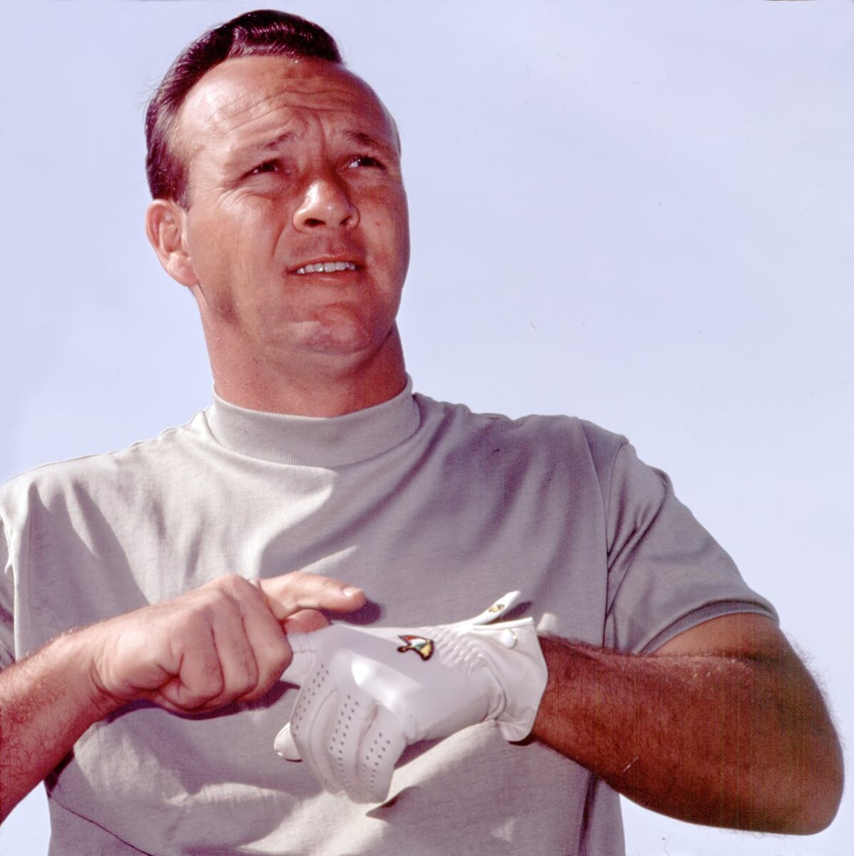 A man in a light shirt puts on a white golf glove, looking into the distance against a clear sky.