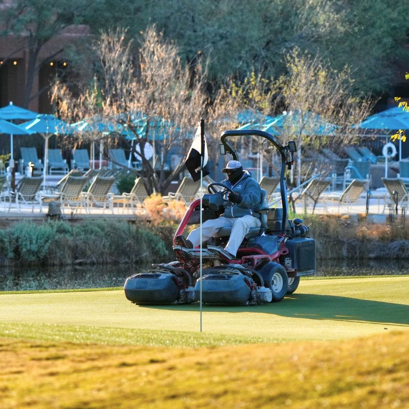 Person mowing a golf course green with a ride-on mower near a pond and poolside lounge chairs.