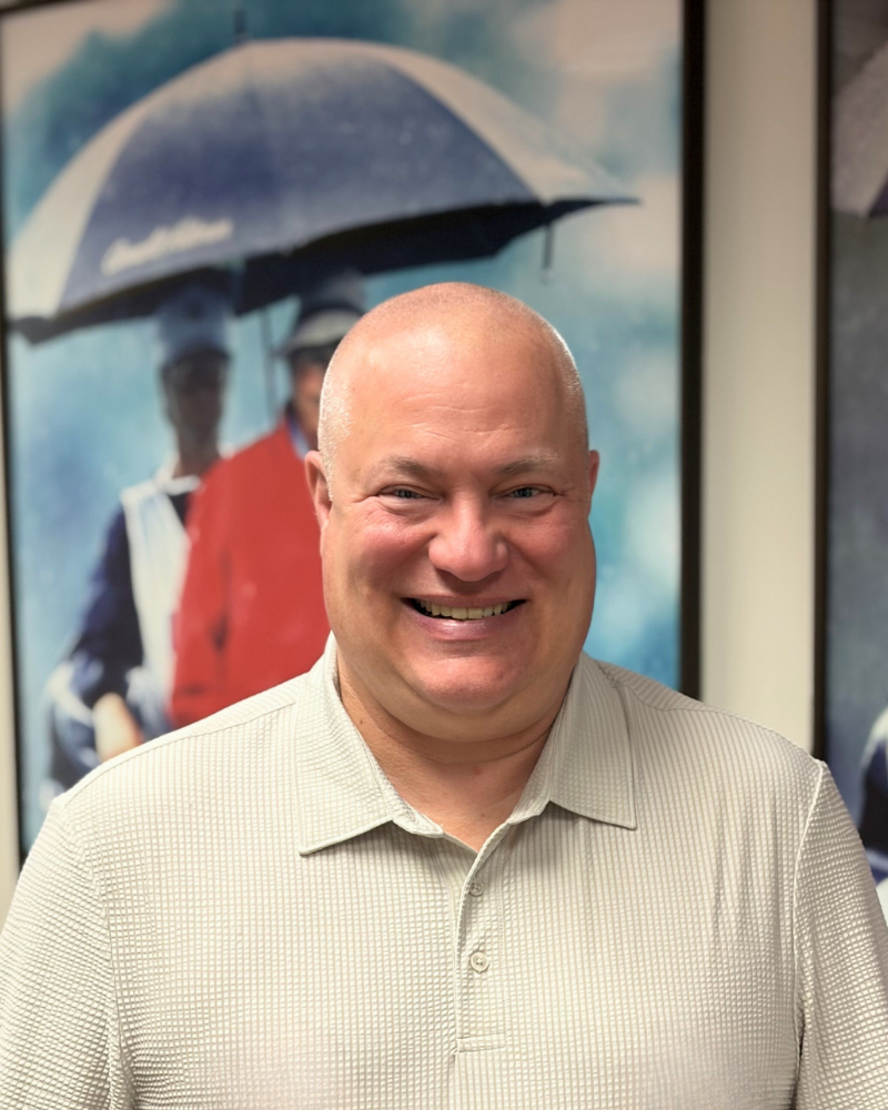 Smiling bald man in a light shirt stands indoors with an umbrella photo in the background.