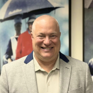 Smiling man in a light gray blazer stands in front of blurred artwork featuring people with umbrellas.