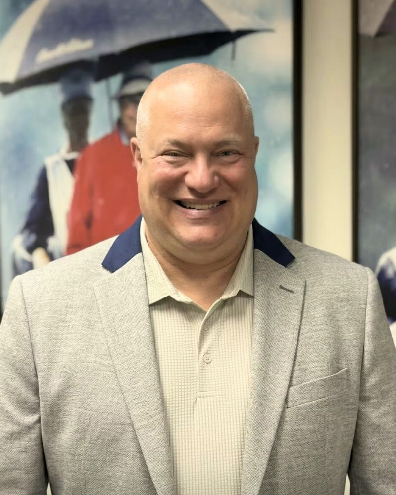 Smiling bald man in a light gray blazer stands in front of blurred artwork featuring people with umbrellas.