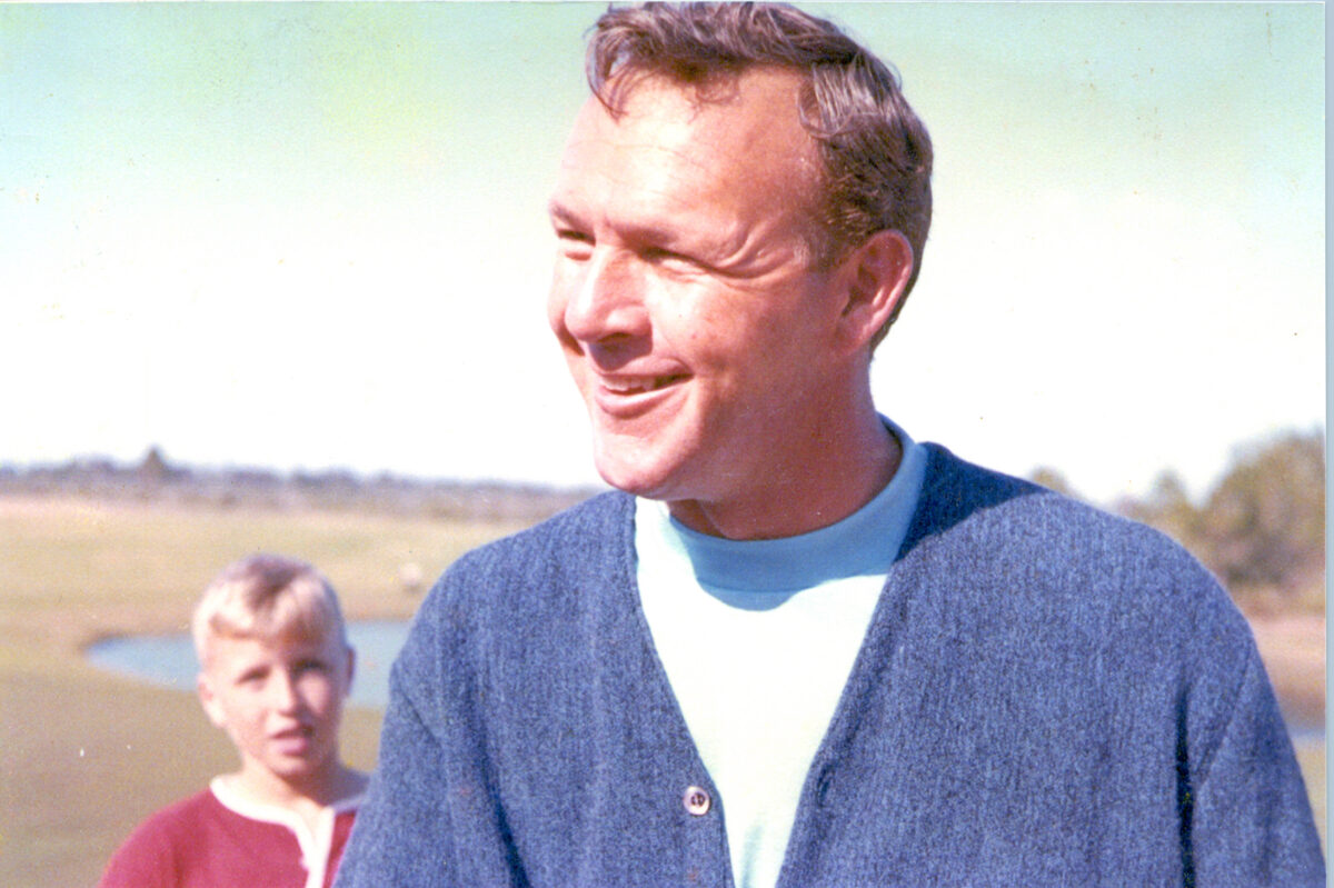 Smiling man in a blue sweater outdoors, with a young boy and grassy field in the background.