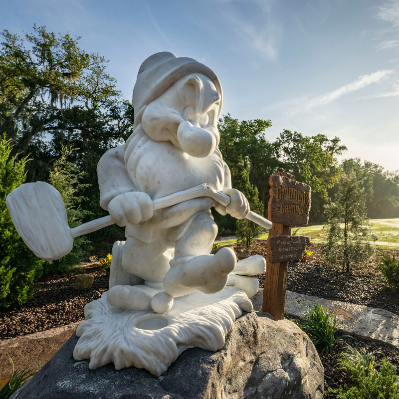Marble statue of Grumpy dwarf holding a golf club, standing on a rock by a wooden sign in a garden setting.