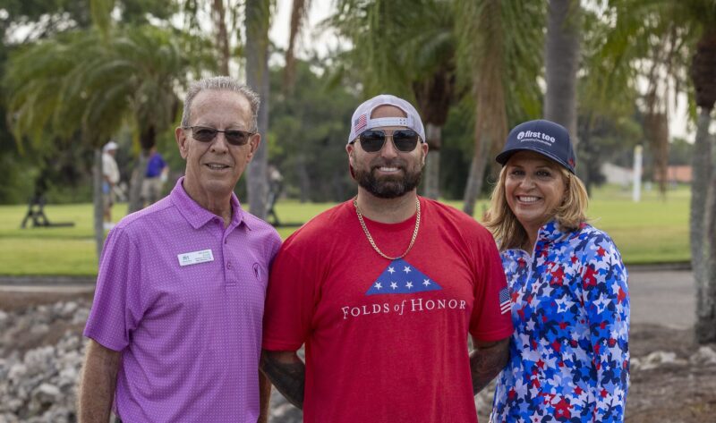 Three people smiling outdoors; one wears a red Folds of Honor shirt, others wear golf attire.