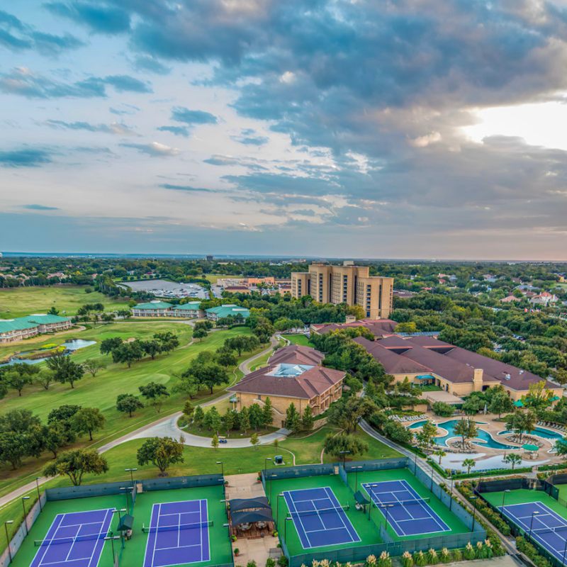 Aerial view of a resort with tennis courts, pools, and buildings surrounded by trees under a cloudy sky.