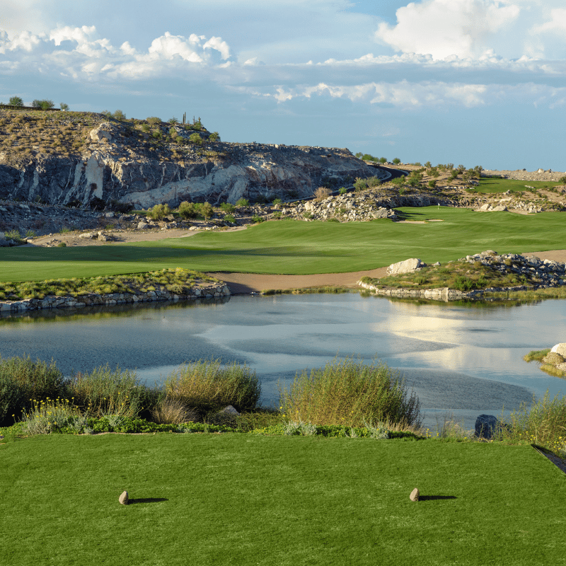A golf course tee box overlooks a water hazard, rocky terrain, and green fairway under a blue sky.