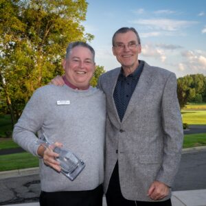 Two smiling men stand outdoors, one holding a glass award, with trees and a golf course in the background.