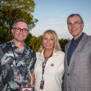 Three smiling adults posing outdoors at sunset, one holding a glass award. Trees and grass in the background.
