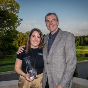 Two people smiling outdoors; the woman holds a glass award. Trees and a fountain are in the background.