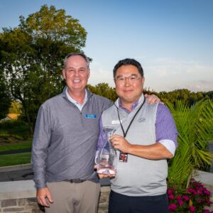 Two men stand outdoors, one holding a glass award, both smiling, trees and golf course in background.