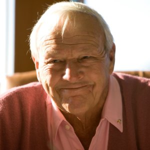 Smiling elderly man in a pink shirt and cardigan, sitting indoors with sunlight on his face.