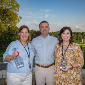 Three adults smiling outdoors, two women holding glass awards, with trees and blue sky in the background.