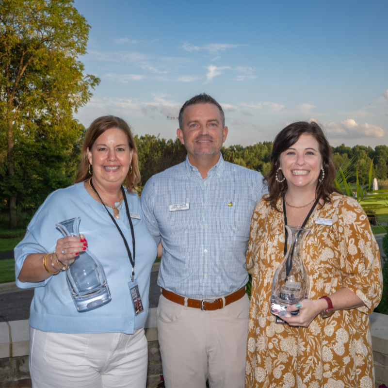 Three adults smiling outdoors, two women holding glass awards, with trees and blue sky in the background.