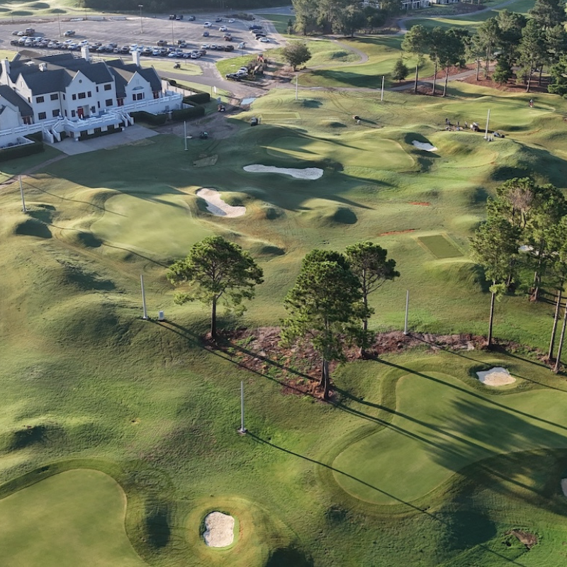 Aerial view of a golf course with sand bunkers, green grass, trees, and a white clubhouse in the background.