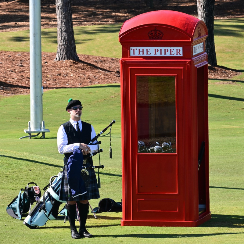 A bagpiper in traditional Scottish dress stands next to a red British-style telephone booth on a golf course.