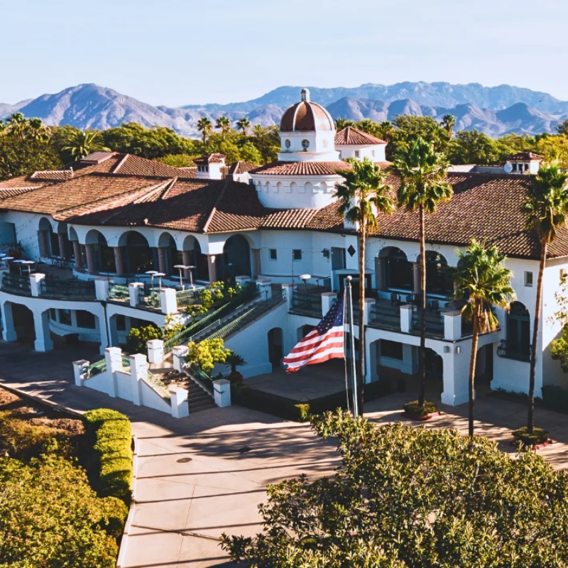 A large Spanish-style building with arches, palm trees, and a U.S. flag, set against mountains.